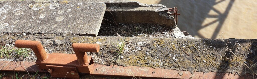 Ruptures. A broken part of a an old harbour wall, in the background sand and the shadow of a bridge.